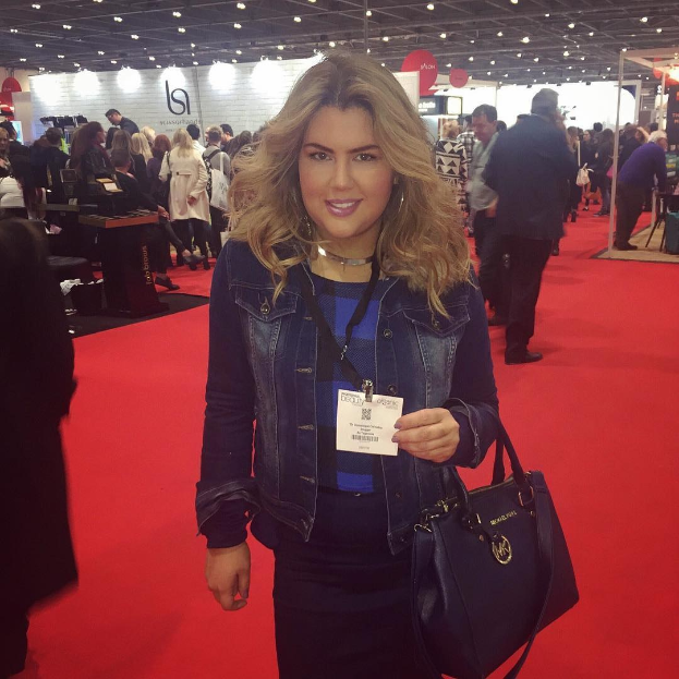 Eine Frau steht auf einem roten Teppich, lächelt und hält eine Handtasche, umgeben von Menschen, Lichtern und Schildern bei einer Modenschau.