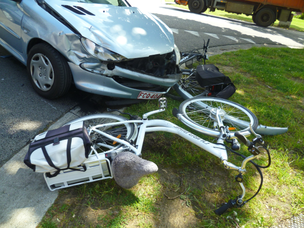 Auto am Straßenrand nach einem Zusammenstoß mit einem Fahrrad, das auf dem Gras liegt, Bäume und klarer blauer Himmel im Hintergrund.