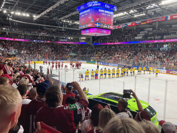 Eishockeyspiel in einer großen Arena mit Spielern auf dem Eis, Zuschauern auf den Rängen, Fotografen, Arena-Beleuchtung und einem Bildschirm mit Text.
