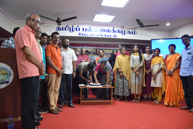 Eine Gruppe von Menschen versammelt sich um einen Tisch mit einem Kuchen in der Nähe eines Podiums mit einem Mikrofon, einer Tafel, einer Flasche, Bannern, einem Bildschirm und Deckenleuchten und -ventilatoren, um die Einweihung einer neuen Schule in Tamil Nadu zu feiern.