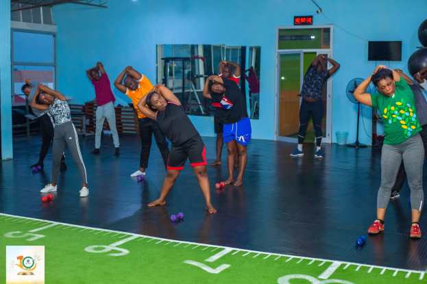 Eine Gruppe von Menschen, die Yoga in einem Fitnessstudio machen, steht auf dem Boden mit Hanteln umgeben von Fitnessgeräten, einem Fernseher, einem Spiegel und Deckenleuchten, mit einem Logo und Text auf der linken Seite.