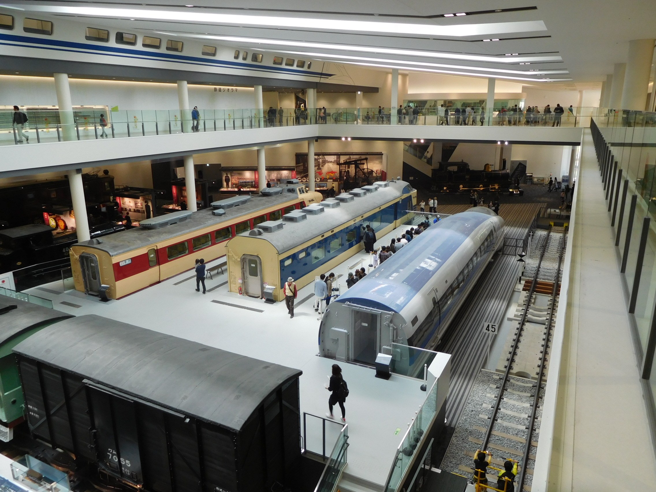 Eine geräumige Museumsausstellung, die verschiedene Züge in unterschiedlichen Größen und Farben zeigt, mit Menschen, die beobachten, und Geländern, Säulen und Deckenleuchten.