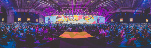 Eine große Zuschauermenge sitzt auf Stühlen einem hell erleuchteten Bühne mit Bildschirmen gegenüber, wo mehrere Menschen während eines Online-Marketing-Events stehen.