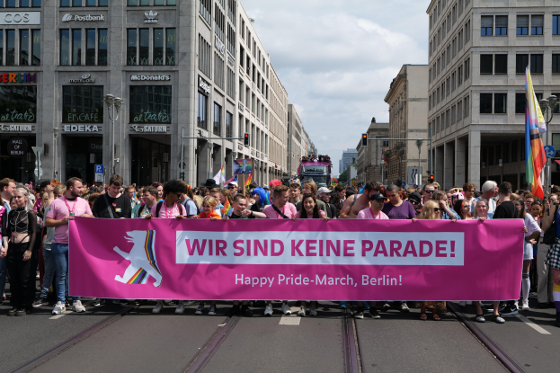 Eine Gruppe von Menschen marschiert eine Straße in Berlin entlang und hält eine pinkfarbene Fahne mit der Aufschrift "Happy Pride March". Die Straße ist von Gebäuden mit Fenstern, Laternenmasten und Verkehrszeichen gesäumt.