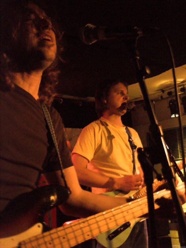 Ein Musikkonzert mit zwei Sängern: einer in einem weißen Hemd, das eine grüne Gitarre hält und ein Mikrofon, und ein anderer in einem schwarzen T-Shirt mit Brille, der eine schwarze Gitarre hält, mit Bühnenbeleuchtung und anderen Instrumenten im Hintergrund.