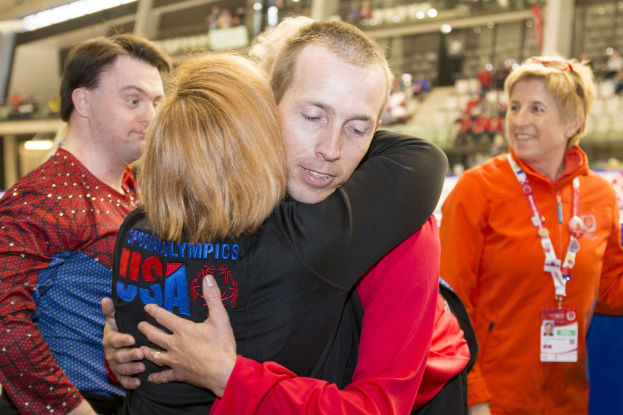 Zwei Menschen umarmen sich in einer herzlichen Umarmung, einer in einem schwarzen und roten Kleid, der andere in einem orangen Kleid mit einem ID-Badge, vor einer unscharfen Menge.