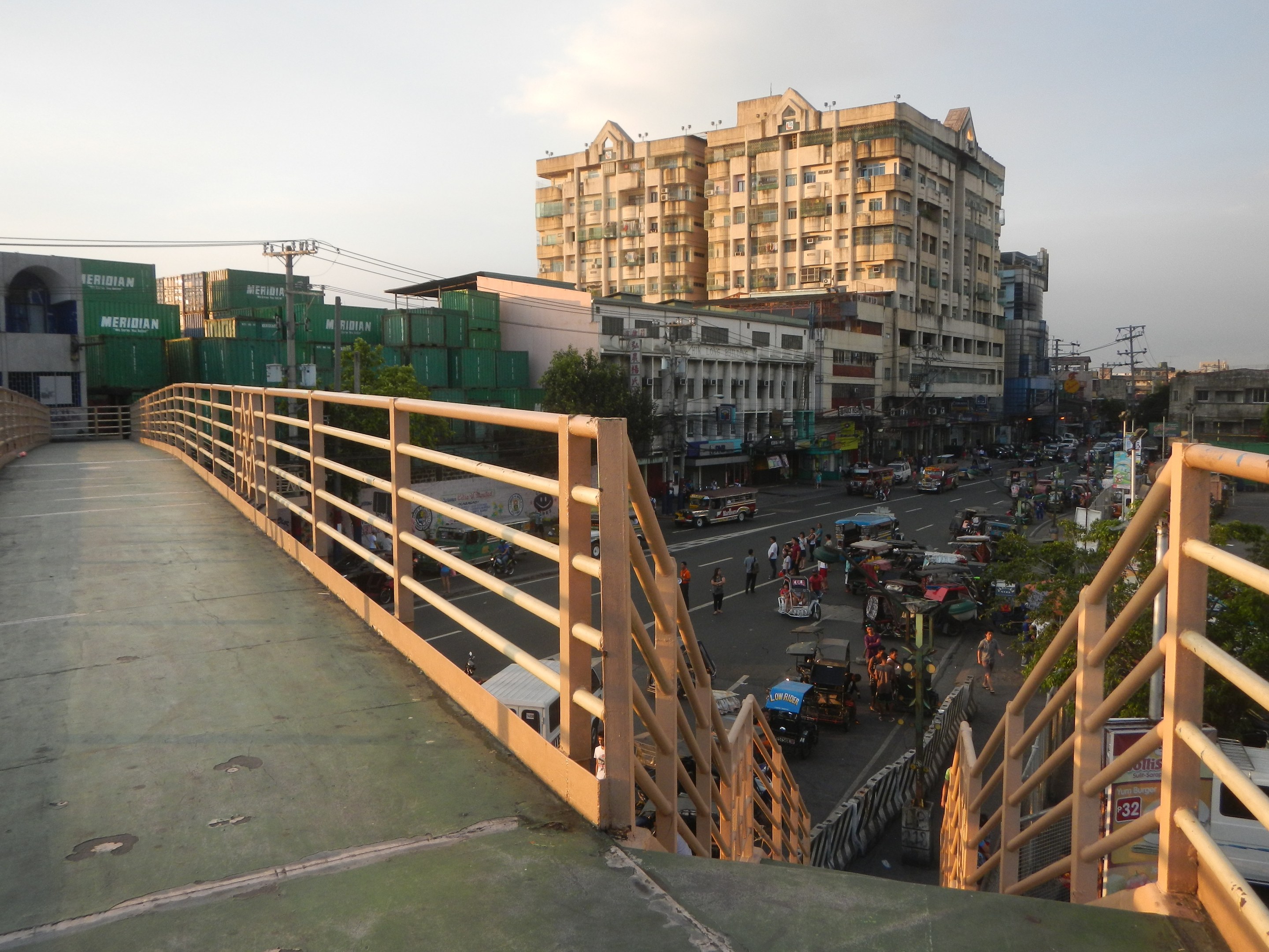 Fußgängerbrücke mit Geländern, die eine befahrene Straße mit vielen Fahrzeugen und Fußgängern überspannt, mit Gebäuden, Bäumen, Strommasten und einem bewölkten Himmel im Hintergrund.