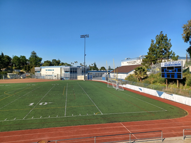 Ein Fußballfeld an der Oakland High School mit einem Torpfosten, umgeben von Gebäuden, Bäumen, Pfählen, Lampen und einem Zaun, mit einem Scoreboard auf der rechten Seite und einem sichtbaren Himmel darüber.