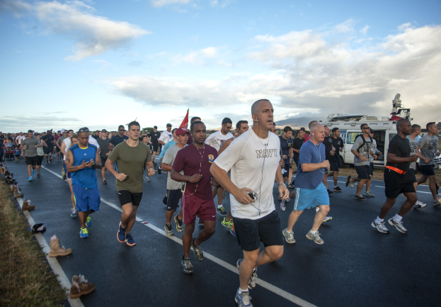 Gruppe von Menschen, die einen Marathon auf einer Straße mit Gras auf beiden Seiten laufen, tragen sportliche Schuhe, mit Fahrzeugen, Bäumen, Hügeln und einem bewölkten Himmel im Hintergrund.