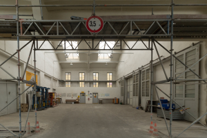 Großer Lagerraum mit ausführlichem Gerüst, Verkehrskegeln, Metallstäben, einem Schild, Maschinen, einer Tür, Fenstern, Wänden, Deckenlampen und Dach.