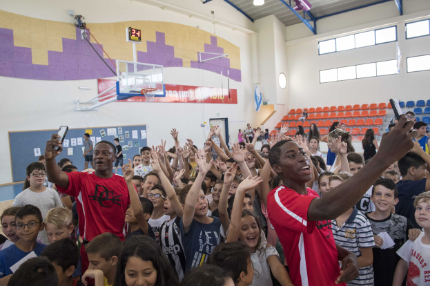 Kinder vor einem Basketballfeld mit Handys stehend, mit einer Tafel, Uhr, Torpfosten, Basketballkorb, Deckenbeleuchtung, Stühlen und Fenstern im Hintergrund.