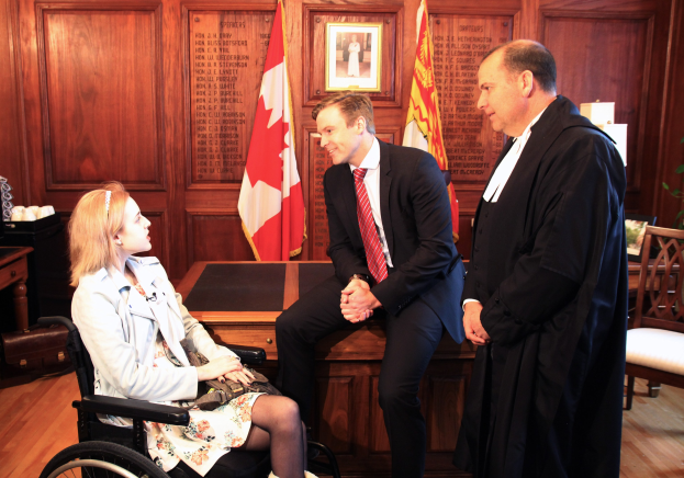 Eine Frau im Rollstuhl spricht mit zwei stehenden Männern in einem Gerichtssaal, mit Flaggen, einem Fotorahmen, einer Pflanze, einem Stuhl und einem Tisch mit verschiedenen Gegenständen im Hintergrund.