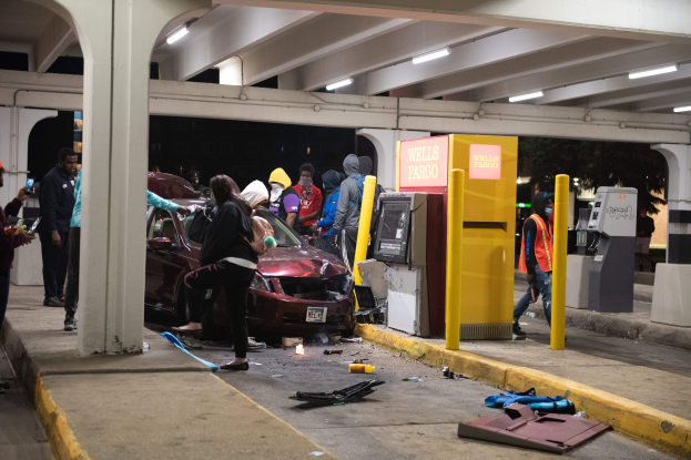 Eine Gruppe von Menschen steht um ein Auto herum, das in eine Tankstelle gekracht ist. Überall liegen Gegenstände verstreut und im Hintergrund sind Tankstellenausrüstung zu sehen.