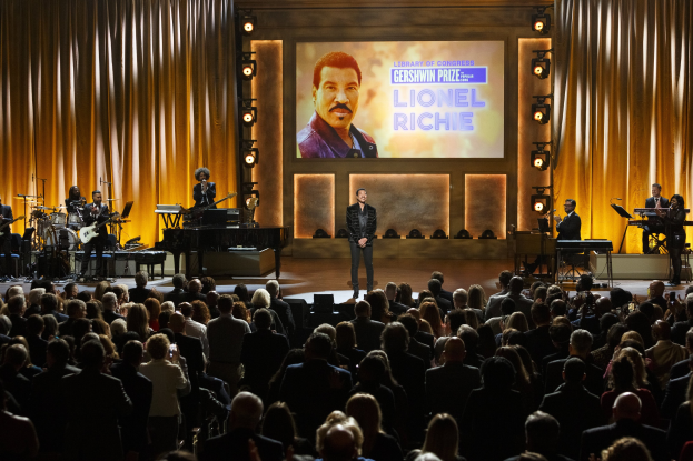 Ein Mann mit einem Mikrofon steht auf einer Bühne vor einem sitzenden Publikum, begleitet von Musikern, die Instrumente spielen, mit einem Display, das Lionel Richie und Text zeigt.