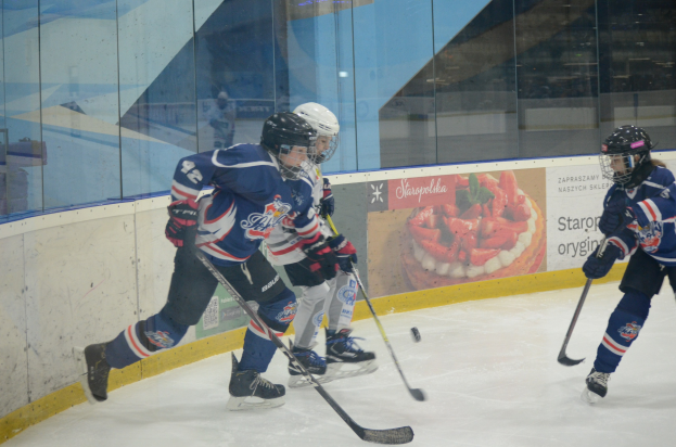 Gruppe junger Menschen beim Eisschlittschuhlaufen auf einer Indoor-Eisbahn, mit Helmen, Sportbekleidung und Hockey-Schlägern, mit einem Plakat im Hintergrund auf einer Glaswand.