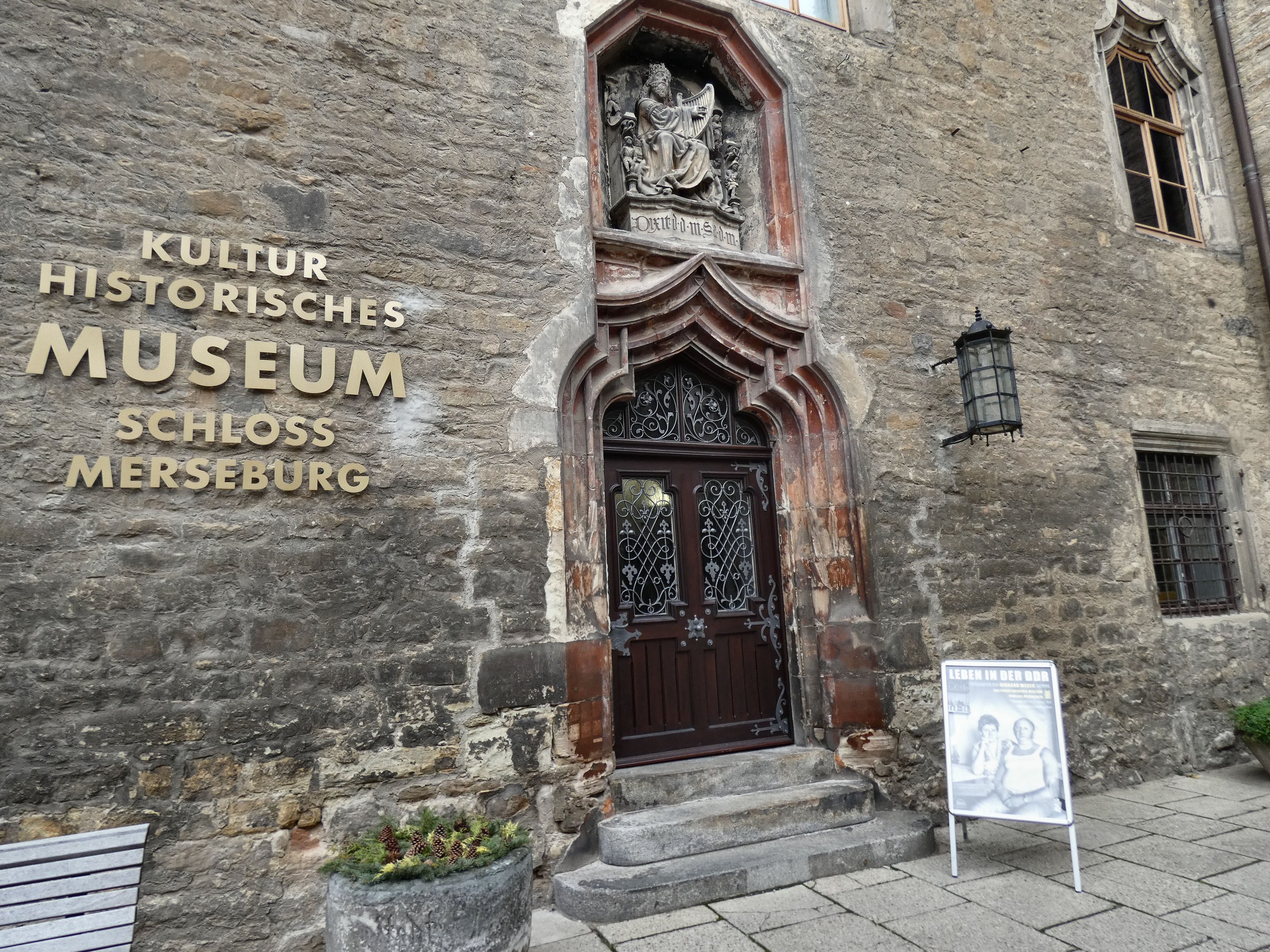Exterior view of the Kultur Historische Museum in Schloss Merseburg, Germany, featuring a building with windows and a door, a signboard, a bench, potted plants, a pipe, and a cloudy sky.