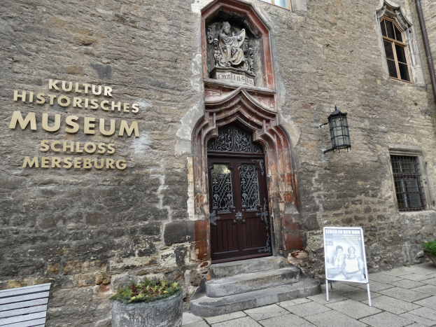 Exterior view of the Kultur Historische Museum in Schloss Merseburg, Germany, featuring a building with windows and a door, a signboard, a bench, potted plants, a pipe, and a cloudy sky.