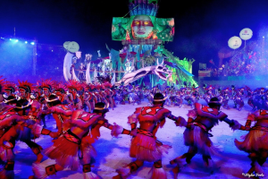 Eine Gruppe von Menschen in bunten Kostümen tanzt auf einer Bühne während des Karnevals 2014 in Rio, vor einem Publikum sitzend, beleuchtet von Lichtern und einem Bildschirm mit einer Person und anderen Gegenständen im Hintergrund.