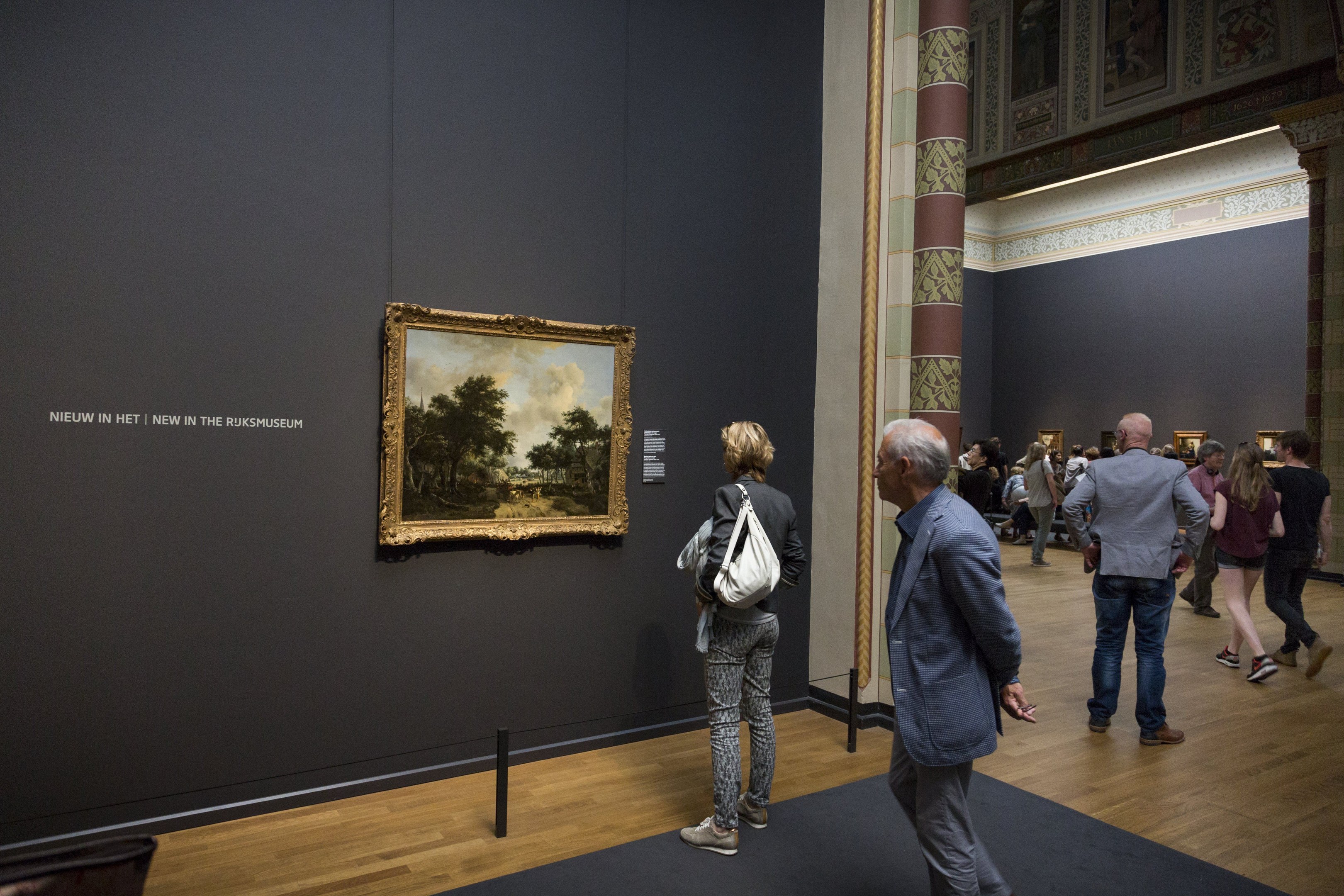 Menschen, die durch ein Museum gehen und ein gerahmtes Gemälde mit einer Absperrung davor betrachten.