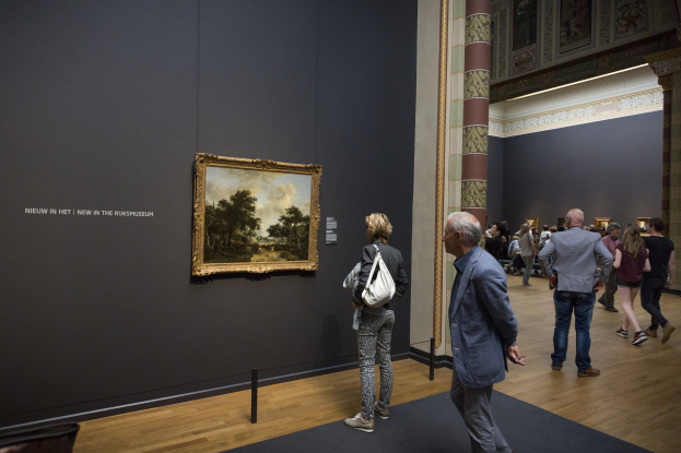 Menschen, die durch ein Museum gehen und ein gerahmtes Gemälde mit einer Absperrung davor betrachten.