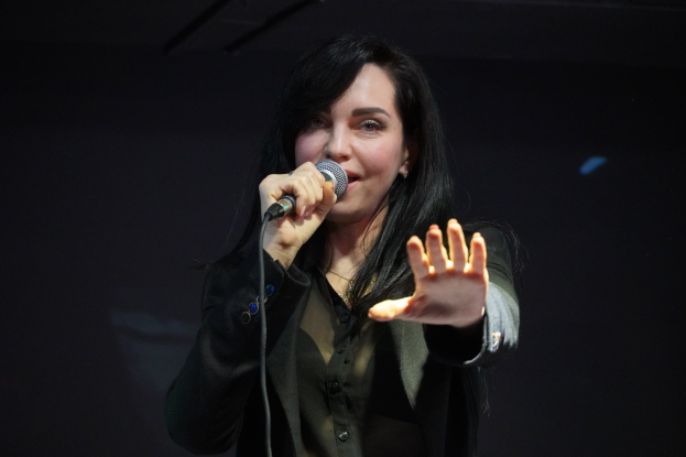 Eine Frau in einem schwarzen Kleid singt in ein Mikrofon auf einer Bühne, ihre Augen geschlossen und ihr Gesichtsausdruck zeigt Emotion und Konzentration, beleuchtet von einem hellen Licht.