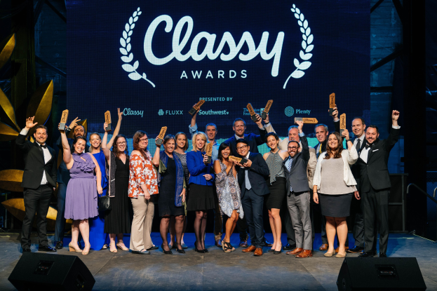 Group of people on a stage holding awards, with speakers below and a screen displaying "Classy Awards 2019" in the background.
