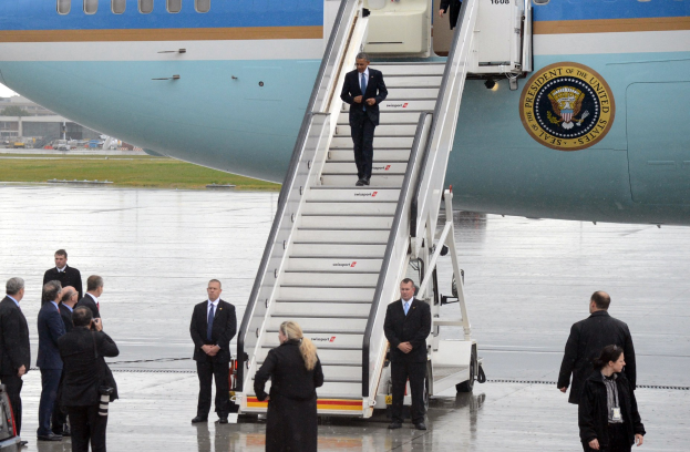 Präsident Obama besteigt Air Force One am Flughafen, umgeben von Mitarbeitern in schwarzen Blazern, weißen Hemden und schwarzen Hosen, mit einem Fahrzeug, Gras, Gebäuden und einem klaren blauen Himmel im Hintergrund.