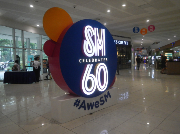 Eine Gruppe von Menschen steht auf einem Einkaufszentrumsboden unter einem "SM feiert 60-jähriges Jubiläum"-Schild, mit einem linksseitigen schwarzen-gekleideten Tisch und Glastüren, Schildtafeln und Deckenlampen im Hintergrund.