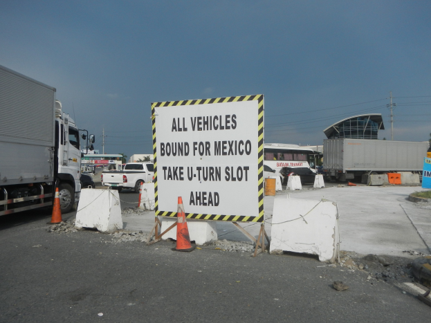Ein Lastwagen auf einem Parkplatz neben einem Schild mit der Aufschrift "Alle Fahrzeuge nach Mexiko". Im Hintergrund sind Gebäude, Bäume, Pfosten und ein klarer blauer Himmel zu sehen.