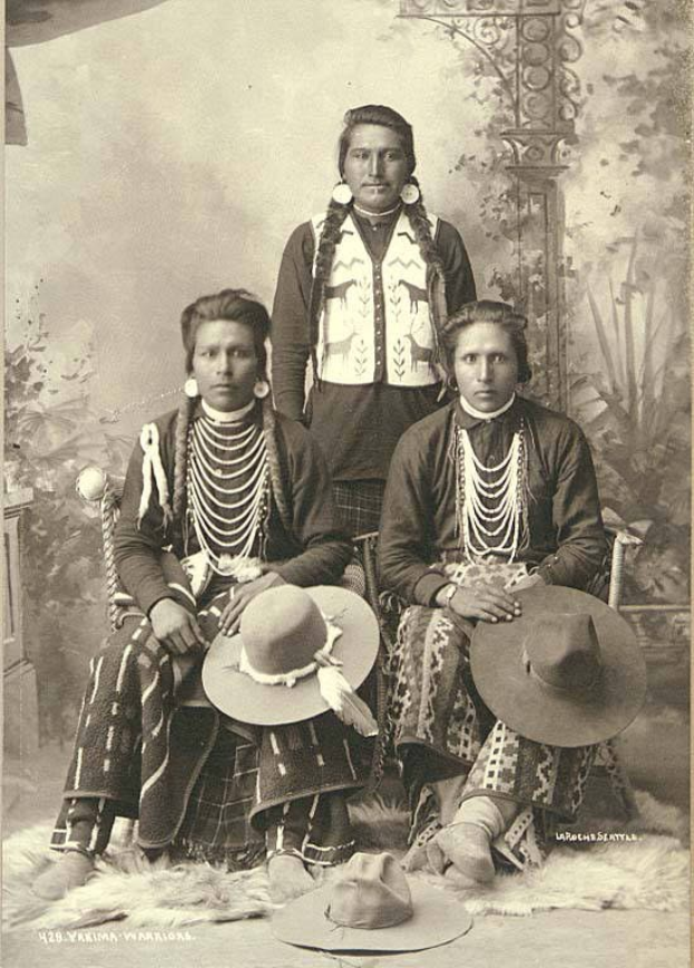 Schwarzes und weißes Foto von drei indigenen Amerikanern, die auf einem Stuhl sitzen, in traditioneller Kleidung und Hüten, mit Pflanzen und einer Wand im Hintergrund und Text unten.
