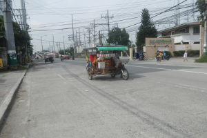 A busy street with parked rickshaws, pedestrians on a sidewalk, electric poles, trees, buildings, a signboard, and visible sky.