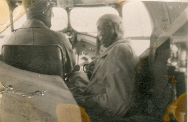 Schwarzes und weißes Foto von zwei Männern in einem Cockpit, einer sitzt auf dem Pilotensitz und trägt eine Uniform, der andere steht neben ihm, beide haben ernste Gesichter.