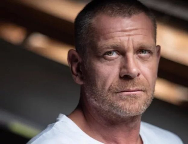 A man in a white t-shirt looking directly at the camera with a blurred background, appearing distressed.