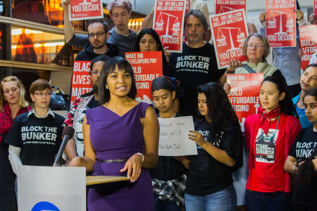 Eine Black-Lives-Matter-Aktivistin spricht an einem Rednerpult mit Mikrofon zu einer Menge mit Schildern, mit Bühnenlicht und Equipment im Hintergrund.