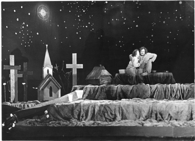 Black and white image of a man and woman embracing on a bed with houses, crosses, and a starry sky in the background.