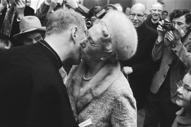 A black-and-white photograph of a man and woman kissing while surrounded by a crowd holding cameras.