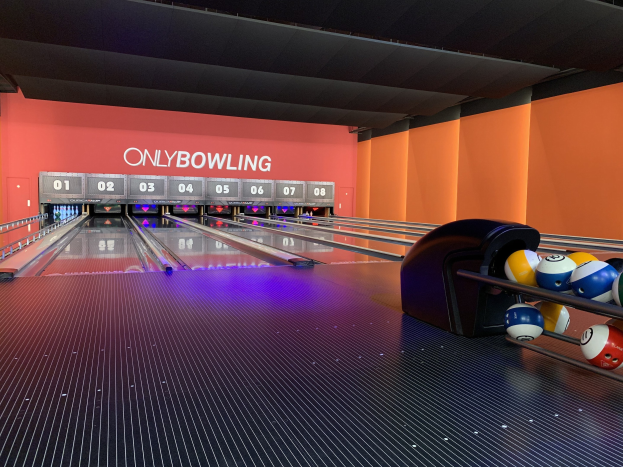 Eine Bowlingbahn mit Bowlingkugeln auf der rechten Seite, Kegel in der Mitte und einer Wand mit Text im Hintergrund, unter einer sichtbaren Decke.
