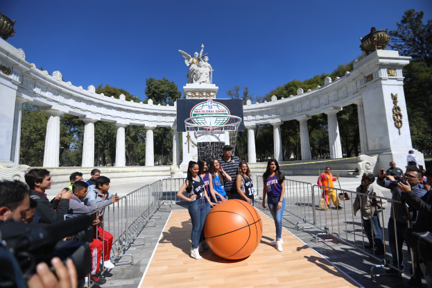 Eine Gruppe von Menschen steht um einen Basketball auf einem Platz mit einem Netz in der Mitte, Geländern auf beiden Seiten und Säulen, Statuen, Bäumen und einem klaren blauen Himmel im Hintergrund, wobei einige Personen Kameras halten.
