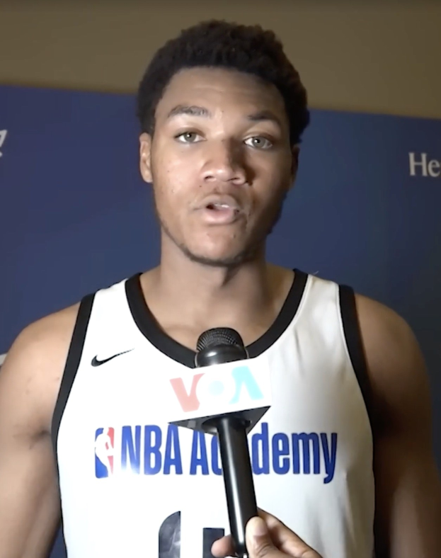 Ein Mann in einem weißen Tanktop hält ein Mikrofon und spricht mit der Medien vor einer Banner mit der Aufschrift "NBA All-Star Game 2019" und einer Wand.