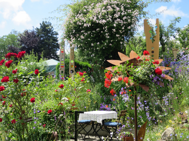 Ein Garten auf dem Chelsea Flower Show mit bunten Blumen und Pflanzen, einem zentralen Tisch mit einer Decke und Bäumen vor einem bewölkten Himmel.