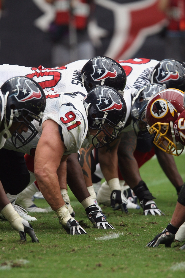 Eine Gruppe von Football-Spielern steht auf dem Feld in einer Reihe, trägt Helme und Handschuhe und hat einen unscharfen Hintergrund.