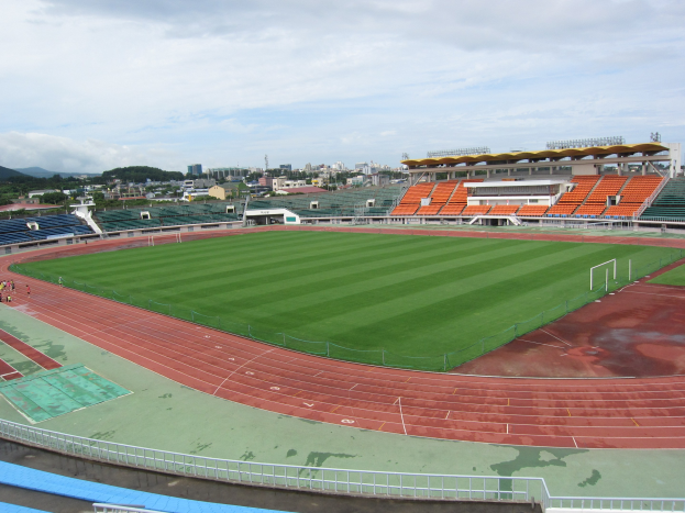 Ein großes Stadion mit einem Fußballfeld, umgeben von Gebäuden, Bäumen, Hügeln und einem klaren blauen Himmel, mit ein paar Menschen auf dem saftig grünen Rasen.