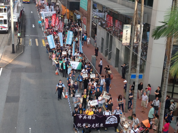 Große Gruppe von Menschen bei einer Protestdemo auf einer Stadtstraße marschierend, mit Schildern und Plakaten, Bäume, Glasgebäude und Fahrzeuge im Hintergrund, was auf eine Demonstration in Hongkong hinweist.