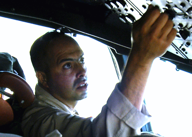 Ein Mann in einem blauen Hemd und schwarzer Mütze sitzt auf dem Fahrersitz eines hellblauen Autos und hält das Lenkrad mit entschlossener Miene fest.