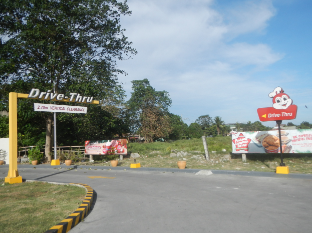 Einfahrt zu einem Drive-in-Restaurant in den Philippinen mit umgebender Vegetation, Beschilderung und einem bewölkten Himmel.