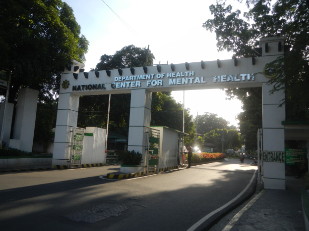 Eintritt zum Nationalen Zentrum für Psychiatrie in Manila, Philippinen, mit einem Tor mit Text, umgeben von Grün, mit einer Straße darunter und Himmel im Hintergrund.