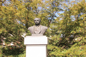 Büste von Wladimir Lenin in Anzug und Krawatte auf einem Sockel, mit Bäumen und einem klaren blauen Himmel im Hintergrund.