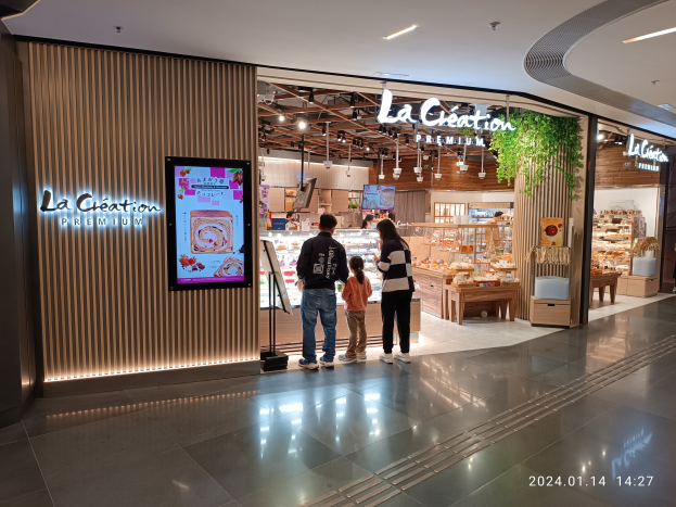 Eine Gruppe von Menschen steht vor dem neu eröffneten La Création Premium Store in einem Einkaufszentrum, mit einem Bildschirm daneben und Essensartikeln im Hintergrund.