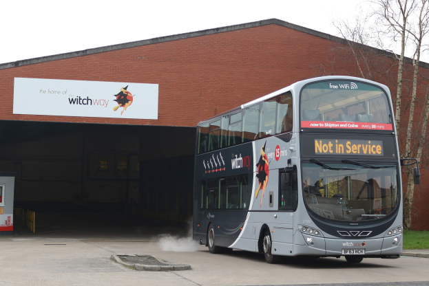 Doppelstockbus mit Rauch vorbei an einem Gebäude fahrend, mit Gras, Bäumen und einem klaren Himmel im Hintergrund; sichtbares Kfz-Kennzeichen am Bus.