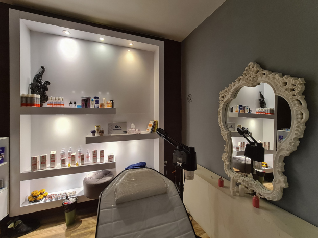 Ein Schönheitssalon mit einem Bett, einem Wandspiegel, Regalen mit Flaschen und Produkten, einem Bodenmülleimer und Deckenleuchten.